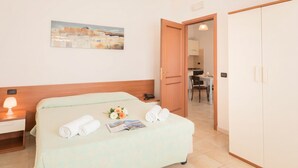 Een hotelkamer met een bed, een nachtkastje met een lamp, een tafeltje met een boek en bloemen, een schilderij aan de muur en uitzicht op de aangrenzende kamer.