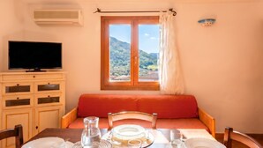 A room with a dining table set for two, a television, a window with a view of hills, and a red cushioned bench.