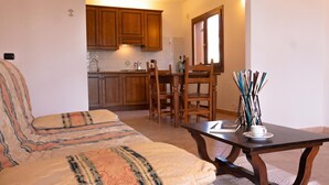 A living room with a sofa, a dining table, and chairs. There is a kitchen area in the background with wooden cabinets and a window. - Cala Viola Trilo 1.5Km From The Beach, Orosei, Italy (Orosei)