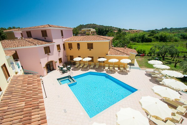 A swimming pool with sun loungers and umbrellas, surrounded by buildings with terracotta roofs.