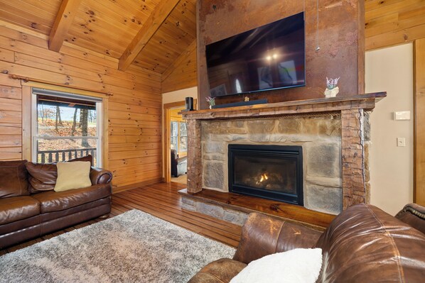 Interior acogedor de una cabaña con chimenea de piedra, muebles de cuero y un televisor de pantalla plana grande montado sobre la chimenea.