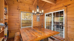 L’intérieur d’un chalet en bois comprend une grande table à manger, un lustre et une vue sur une terrasse extérieure, accessible par une porte vitrée.