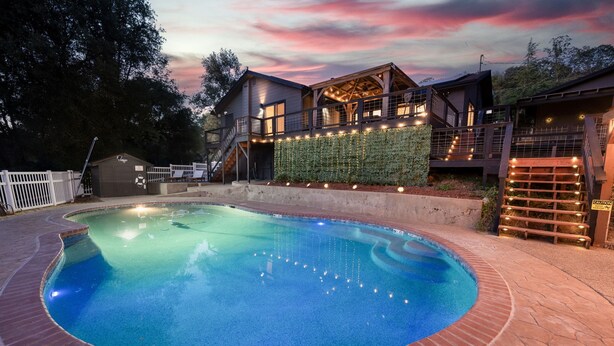 A swimming pool with a brick border, a wooden deck, and a house with a glass facade and outdoor lighting. - Yosemite zen, Pool, Hot tub, Private suite and Views! (oakhurst)