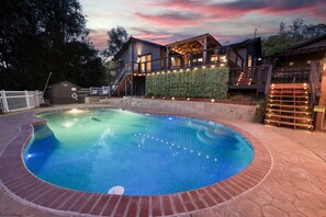 Una piscina con borde de ladrillo, una terraza de madera y una casa con fachada de vidrio e iluminación exterior.