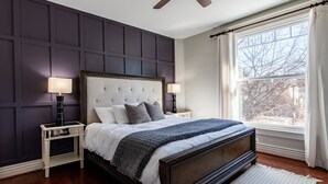 A bedroom with a dark wood bed, white bedding, two bedside tables with lamps, a large window with curtains, and a ceiling fan.