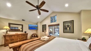 A bedroom with a bed, a wooden bench, a ceiling fan, a dresser, a chair, a window with a view, and framed pictures on the wall.