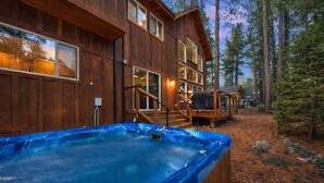 A wooden cabin with a hot tub, a deck, and a view of the surrounding forest.