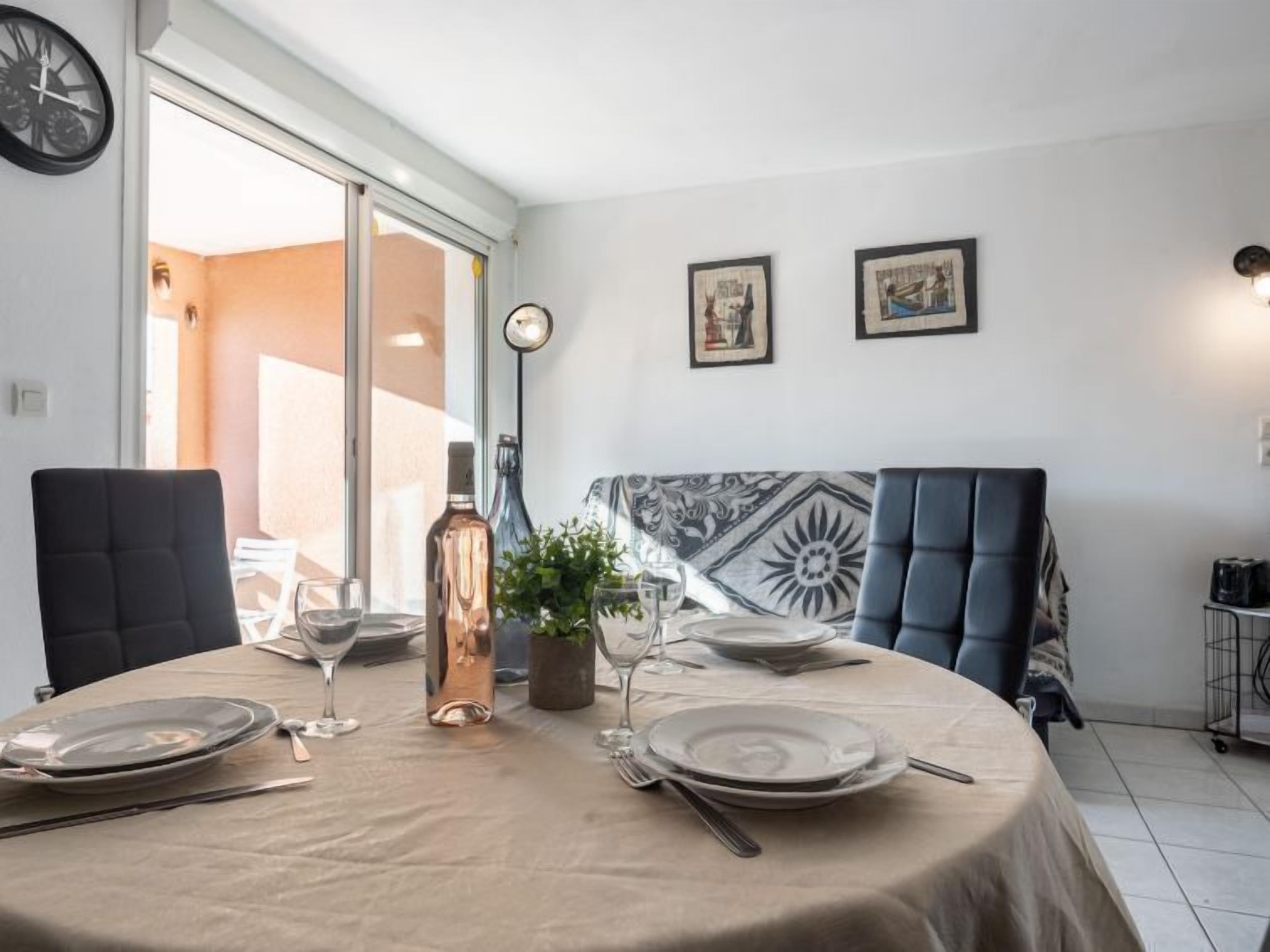 Uma sala de jantar com uma mesa posta para quatro pessoas, um relógio na parede e duas imagens emolduradas.
