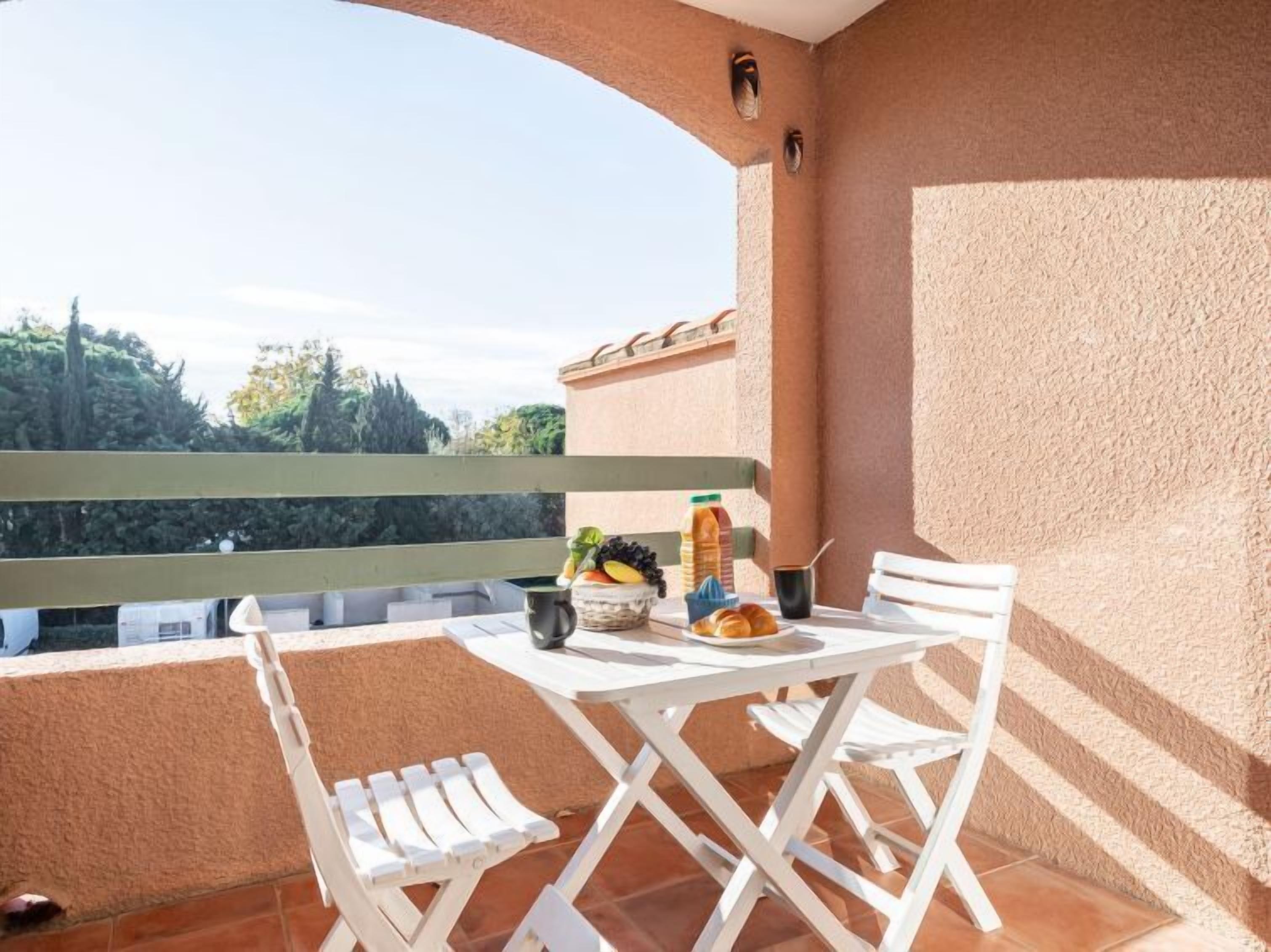 Un balcón con mobiliario blanco, una mesa con comida y bebidas, y vistas a árboles y edificios.