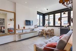 Uma sala de estar moderna com um sofá, uma mesa de jantar e uma televisão. Há uma janela grande com vista para edifícios.