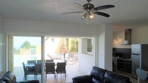 A modern living room with a ceiling fan, a black leather sofa, a dining area with a table and chairs, and a kitchen area with a refrigerator and stove.