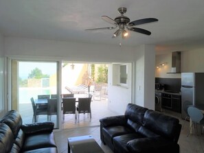 Uma sala de estar moderna com ventilador de teto, um sofá de couro preto, uma área de refeições com mesa e cadeiras, e uma cozinha com geladeira e fogão.