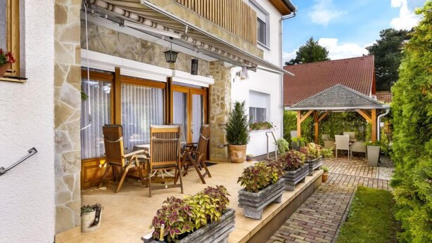 Une maison moderne avec une façade en pierre, un patio couvert avec des meubles d’extérieur et un jardin agrémenté de plantes en pot.