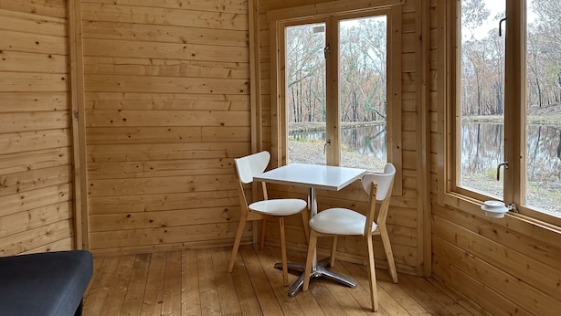 Interior de una cabaña de madera con una mesa de comedor y sillas, ventanas amplias y vistas a un bosque y al agua.