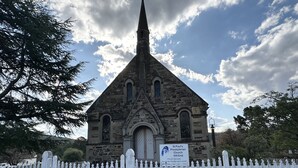 Una iglesia de piedra con un campanario puntiagudo, una valla de piquetes blancos y un cartel que indica los horarios de misa.