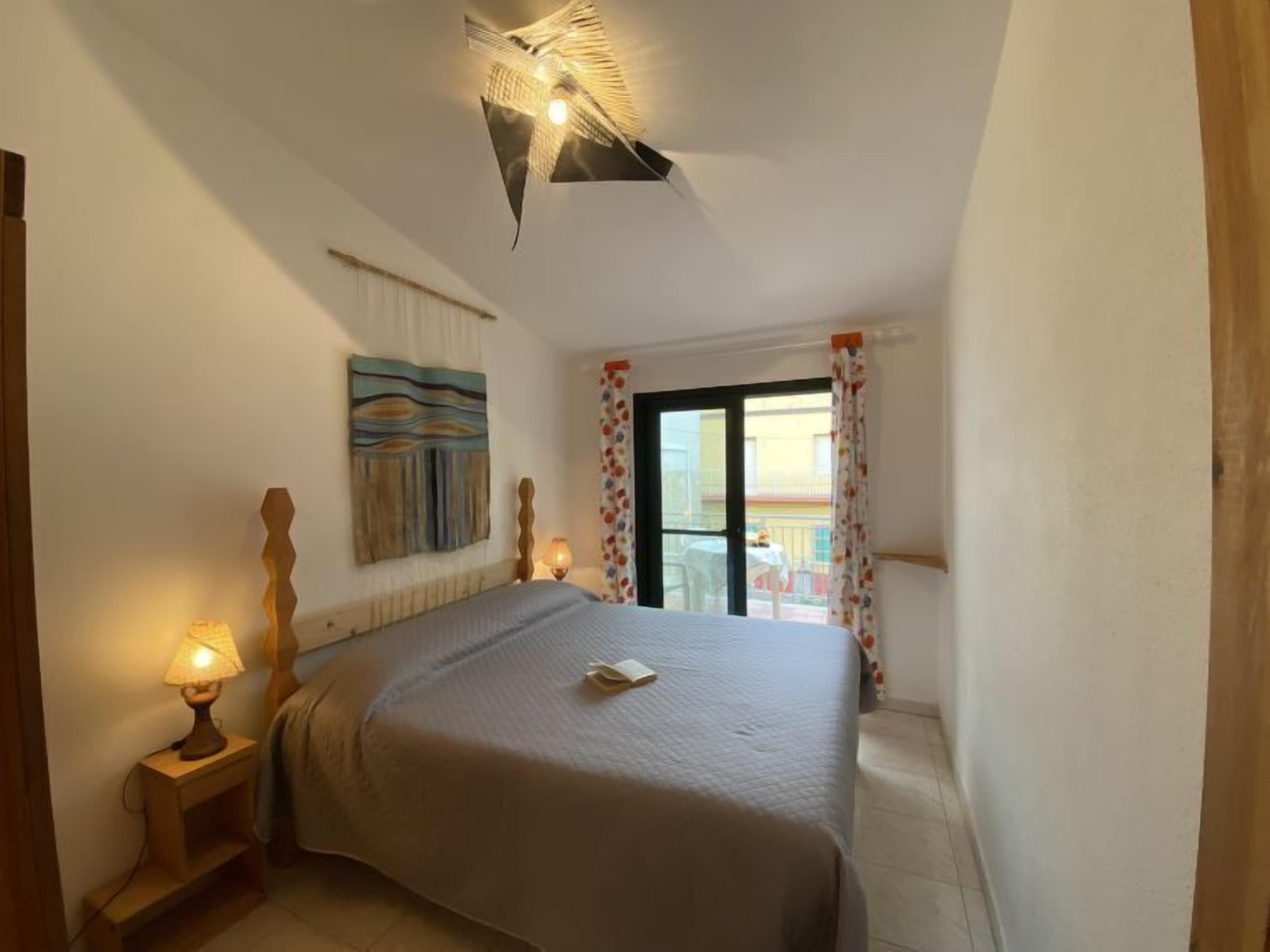 Un dormitorio con cama, mesita de noche con lámpara, lámpara de techo, ventana con cortinas y un cuadro en la pared.