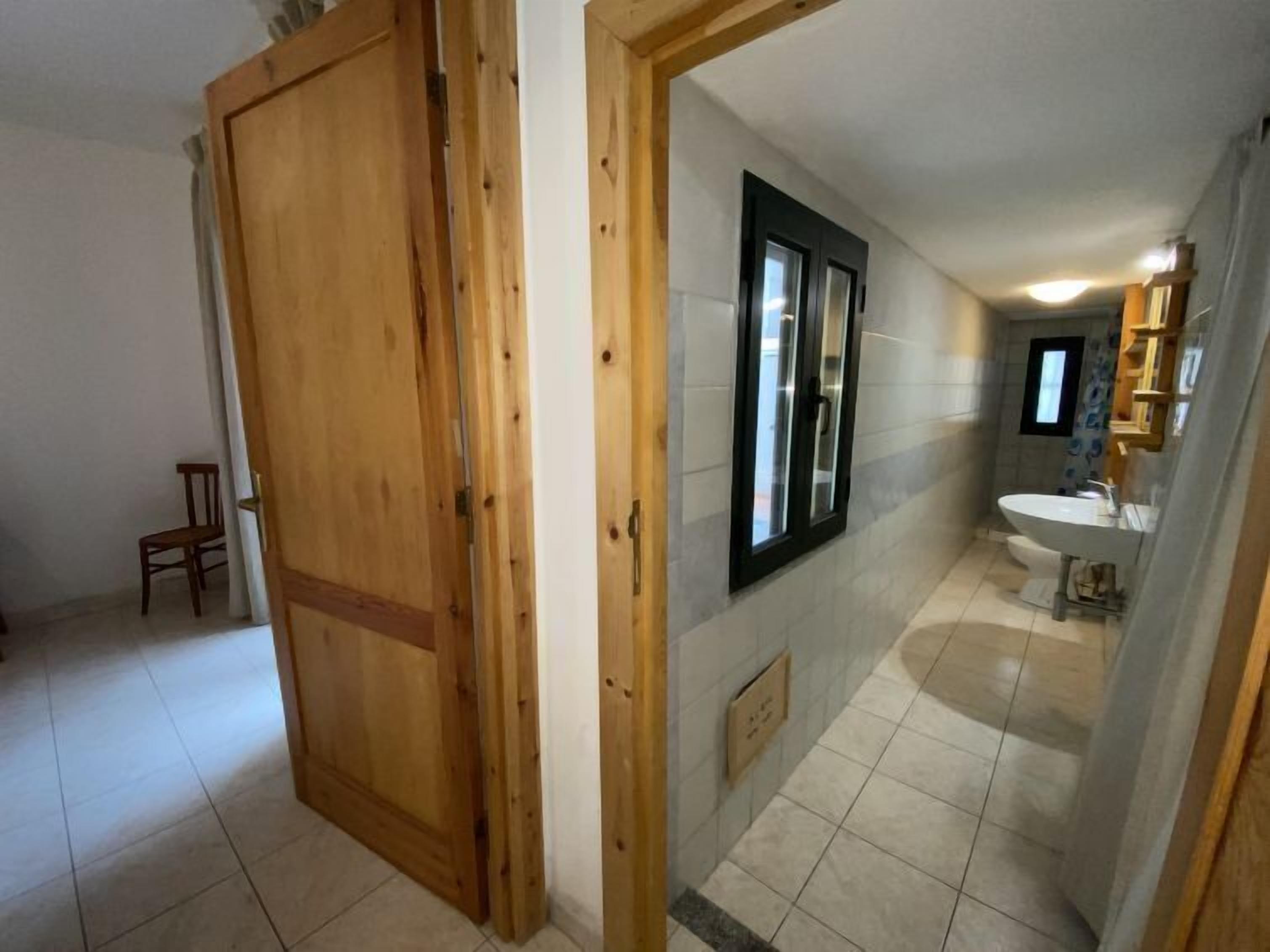 Un pasillo con una puerta de madera, una silla pequeña y un baño con lavamanos y espejo.