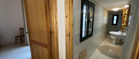Un pasillo con una puerta de madera, una silla pequeña y un baño con lavamanos y espejo.