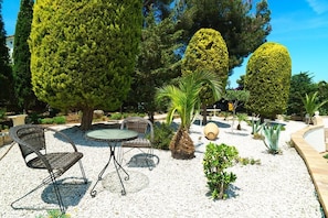 Relax in a serene garden setting with a round table and two wicker chairs surrounded by lush greenery.