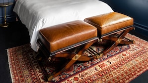 Un dormitorio con una cama grande, dos otomanes de cuero marrón, un tapete estampado y una ventana con vistas.