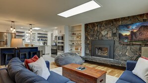 A modern living room with a stone fireplace, blue sofas, and a kitchen in the background. - Wonderful Updated Single Level Retreat w/ Backyard Oasis, Pet Friendly, A/C, Hot Tub, Private Patio (Bend)
