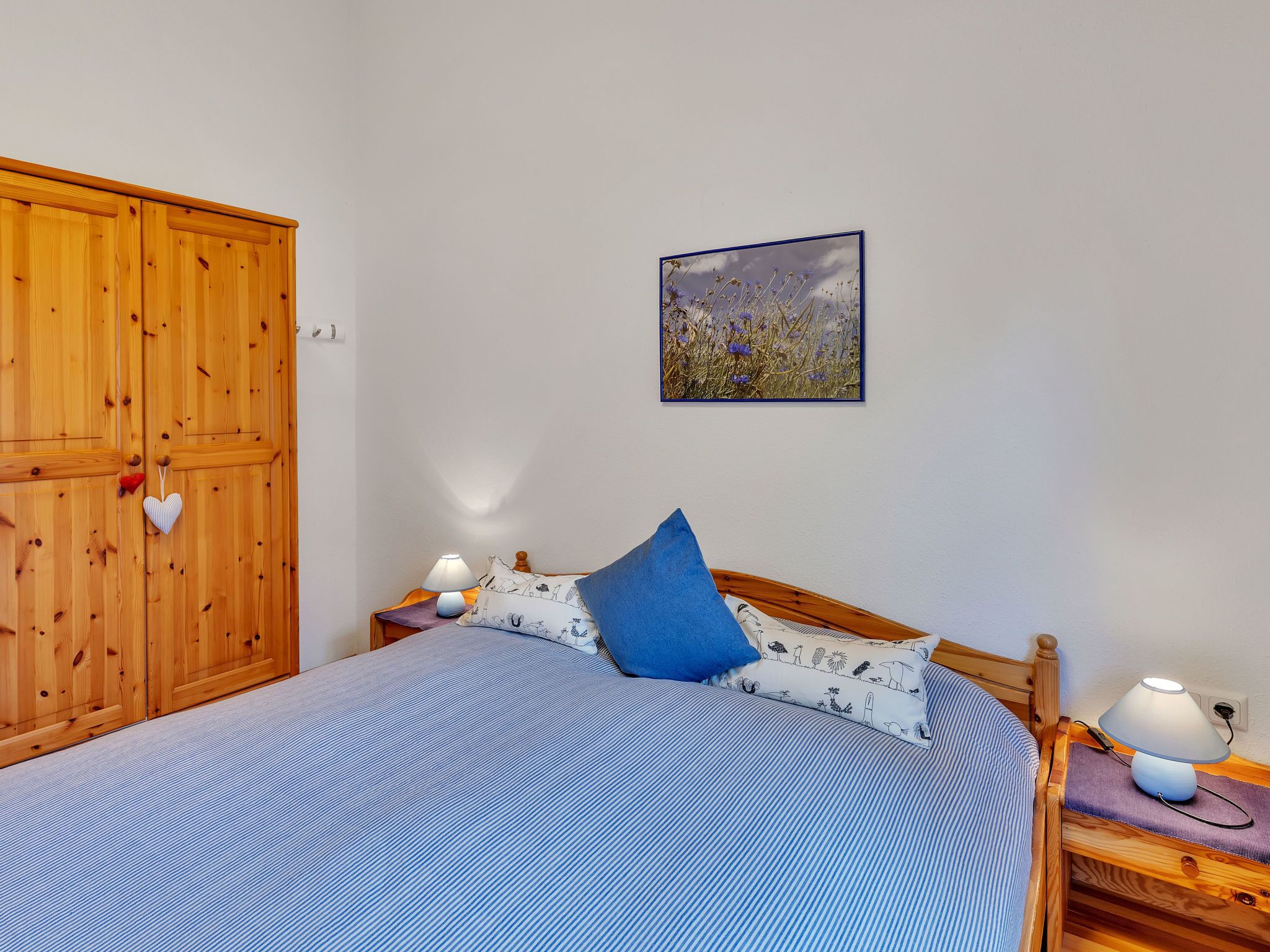 Un dormitorio con una cama, dos mesitas de noche con lámparas, un armario de madera, un cuadro en la pared y un adorno con forma de corazón en el armario.