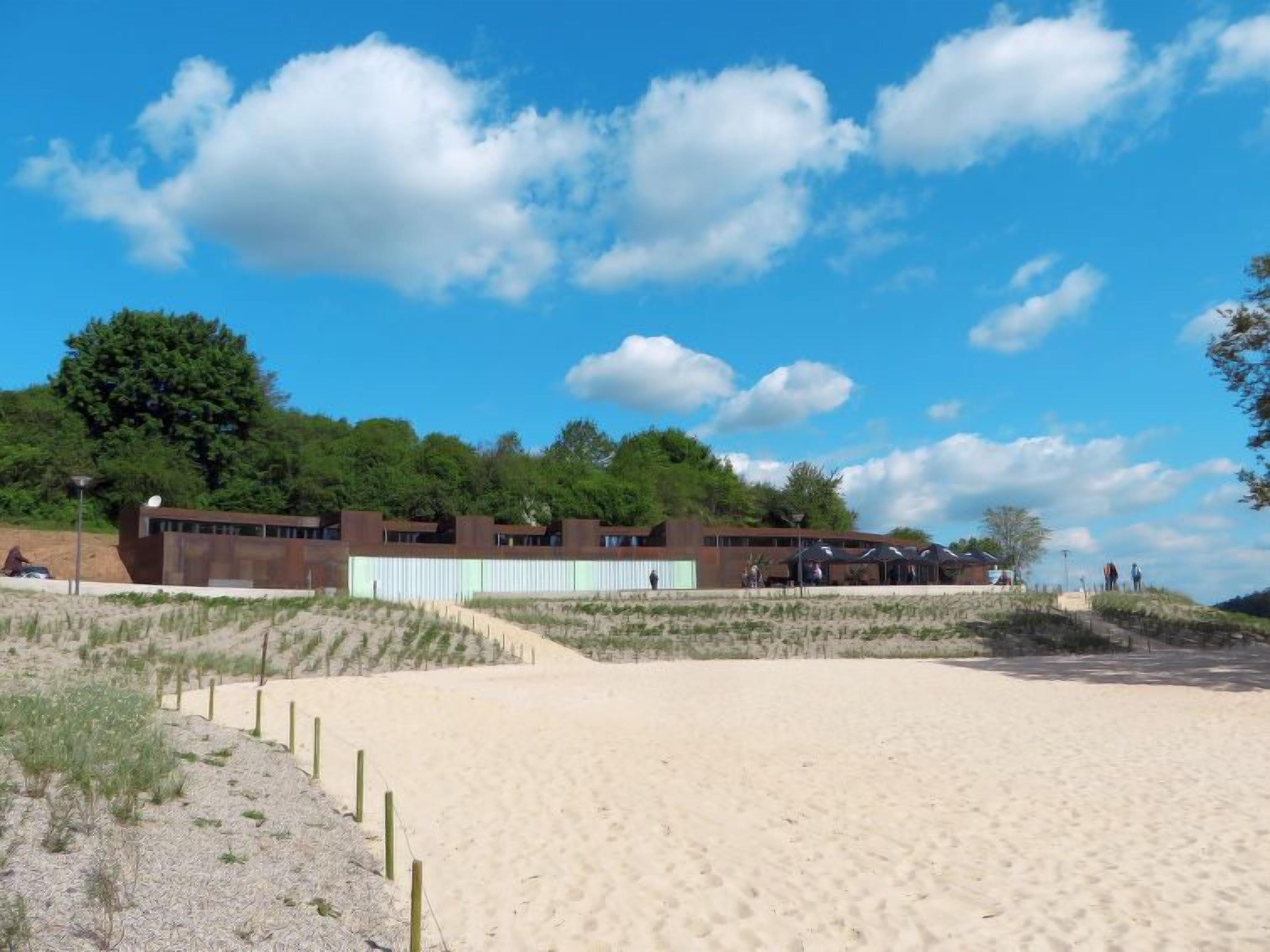 A modern building with a flat roof and large windows, situated on a sandy beach with a clear sky.
