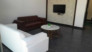 A modern living room with a white sofa, a brown leather couch, and a round coffee table with a plant. A flat-screen TV is mounted on a brick wall. - Yuvraj Palace Inn (Mysore)
