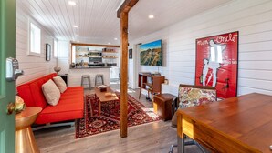 A cozy living room with a red sofa, wooden coffee table, and a red rug. A kitchen with white cabinets and a dining area with a wooden table are visible in the background.