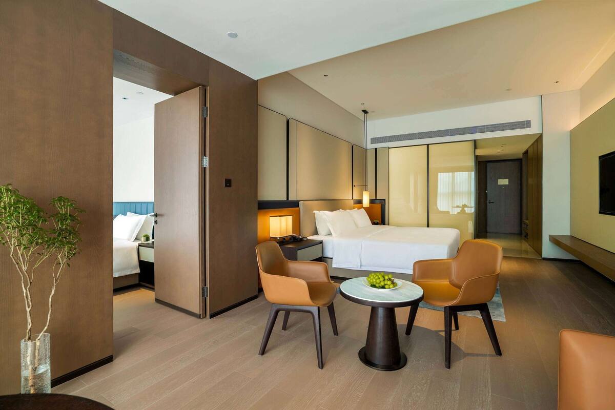 Ein modernes Hotelzimmer mit einem Bett, zwei Sesseln, einem kleinen Tisch mit Obst, einer Topfpflanze und einem großen Spiegel.