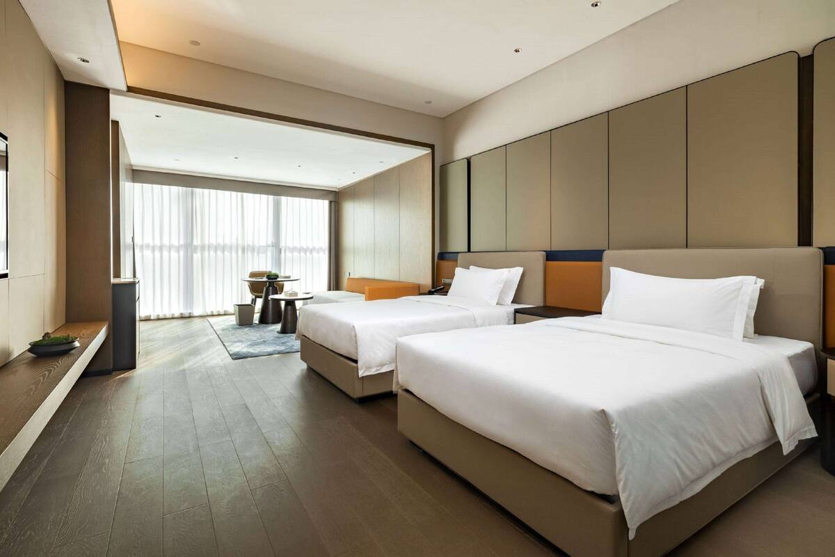 Ein Hotelzimmer mit zwei Betten, einem kleinen Tisch und Blick ins Freie durch große Fenster.