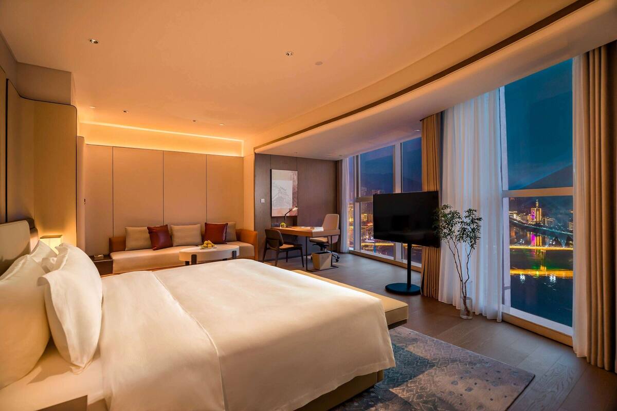 Ein modernes Hotelzimmer mit einem großen Bett, einem Schreibtisch, einem Sessel, einem Fernseher und Blick auf eine nächtliche Stadtlandschaft.