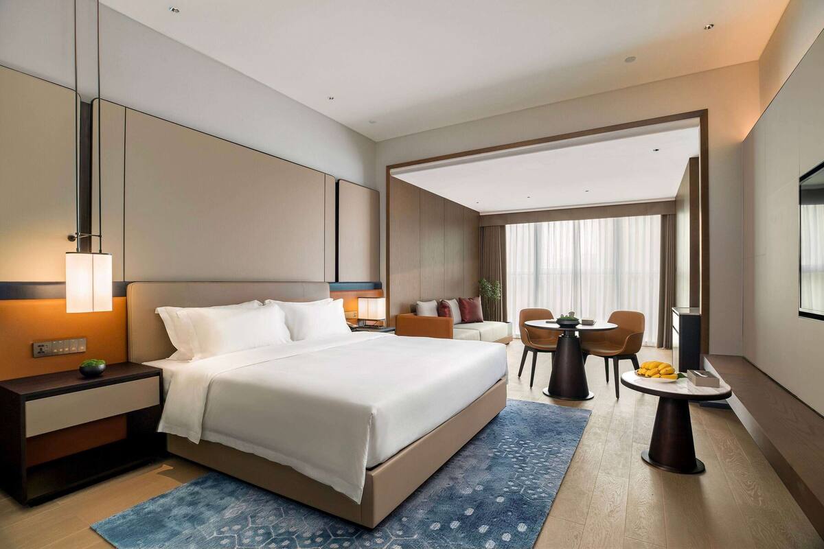 Ein modernes Hotelzimmer mit einem großen Bett, Nachttisch und Lampe. Es gibt einen Essbereich mit Tisch und Stühlen sowie einen Blick auf die Stadt durch große Fenster.