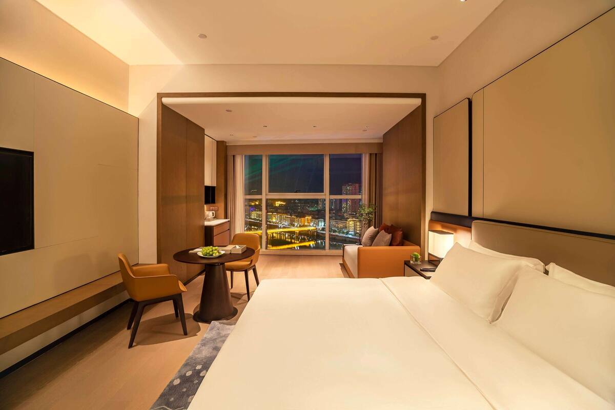 Ein modernes Hotelzimmer mit einem großen Bett, einer Sitzecke mit Tisch und Blick auf eine nächtliche Skyline.