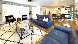 Un salon spacieux avec un canapé bleu, un divan en cuir noir et un tapis à motifs. Au fond, on aperçoit un coin repas avec une table et des chaises.