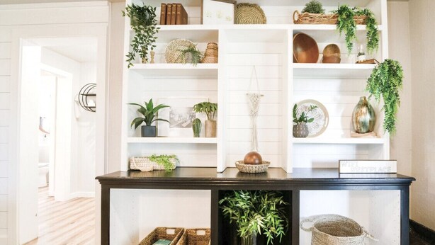 Un comptoir d’accueil avec un revêtement en bois foncé, des étagères blanches au-dessus où sont disposés divers objets décoratifs et des plantes, et une étagère inférieure avec des paniers et une plante en pot.