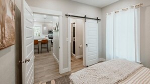Une chambre avec un grand lit, une porte coulissante donnant accès à une salle de bain, et une vue sur la cuisine et l’espace repas.