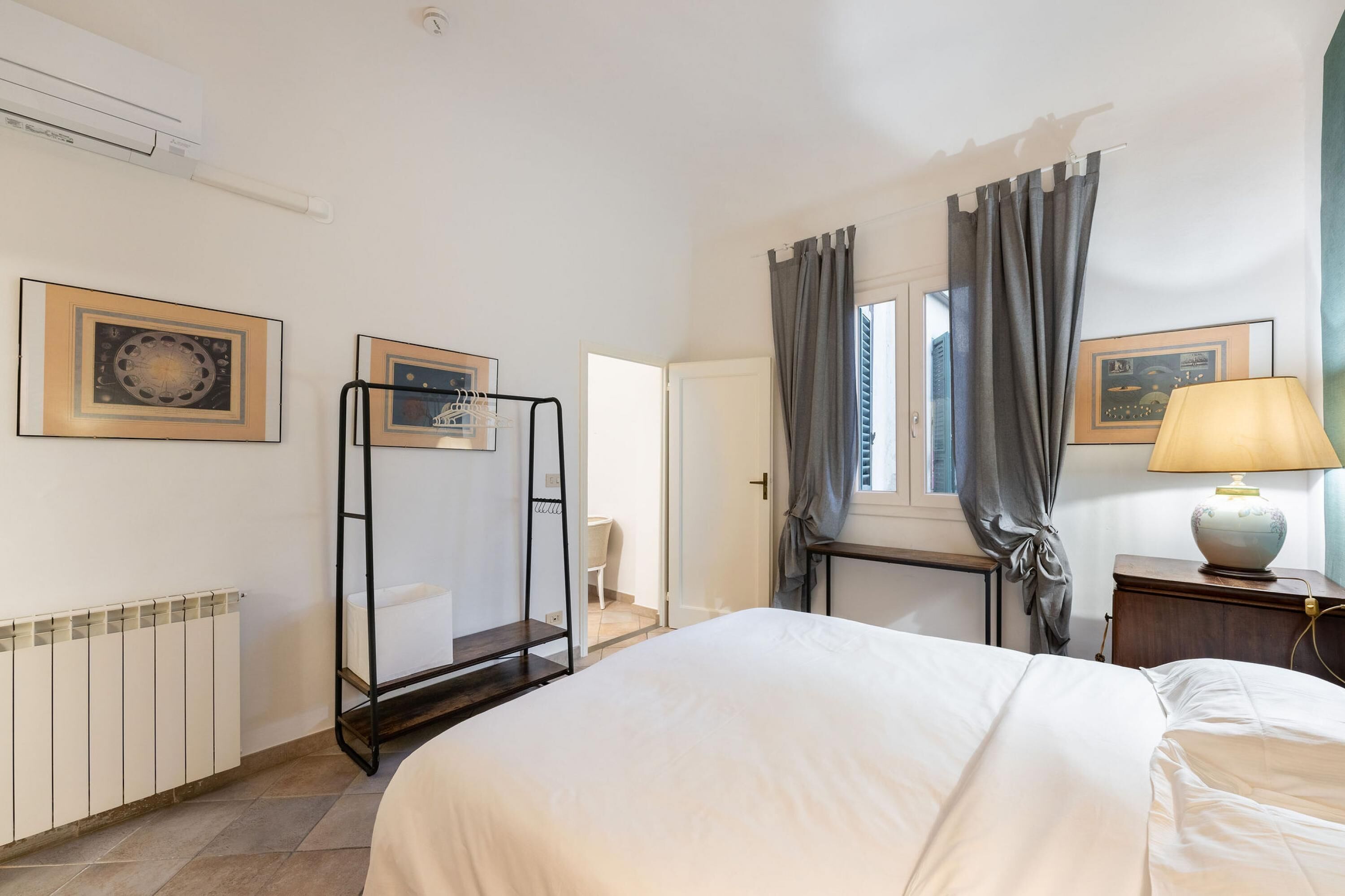 Habitación de hotel con cama, radiador, ventana con cortinas, mesita de noche con lámpara y estante con marcos.