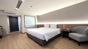Una habitación de hotel moderna con una cama grande, mesitas de noche, una silla y un amplio ventanal con cortinas.