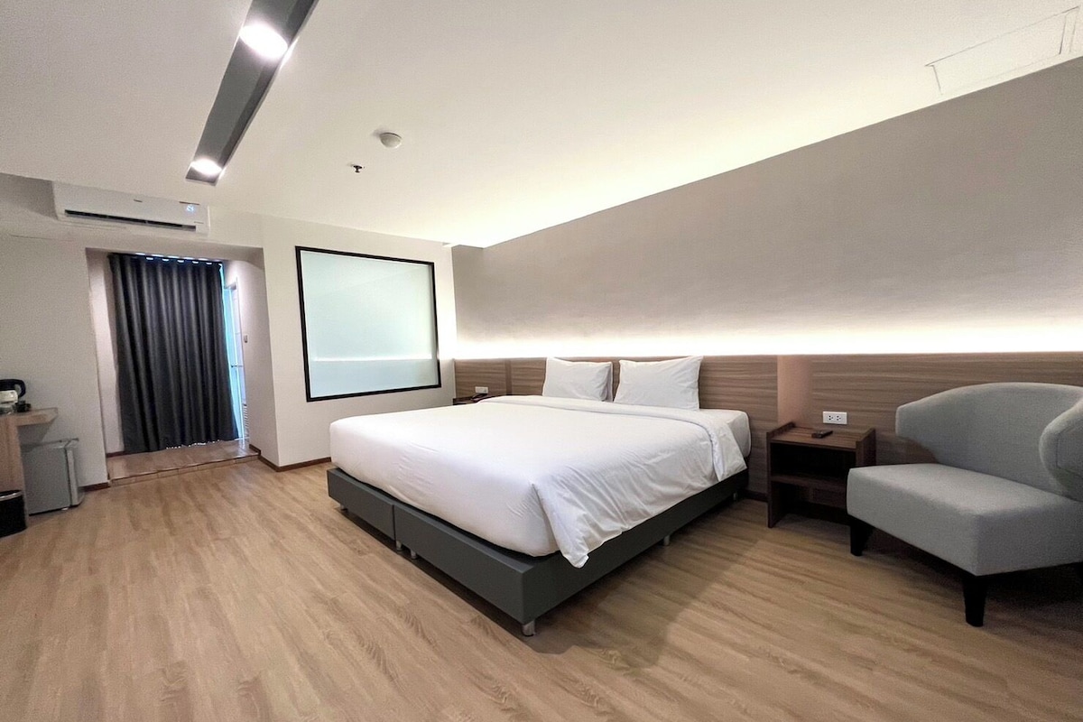 Una habitación de hotel moderna con una cama grande, mesitas de noche, una silla y un amplio ventanal con cortinas.