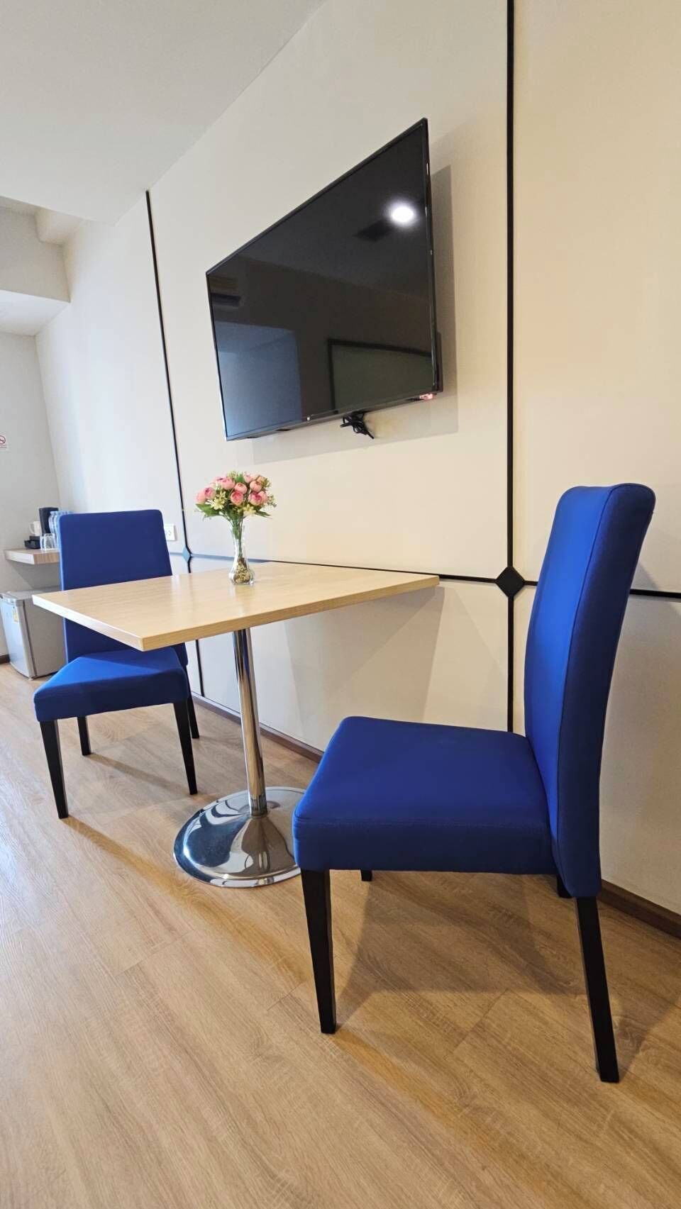 A modern room with a wooden table, two blue chairs, a flat-screen TV mounted on the wall, and a vase with pink flowers on the table.