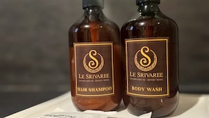 Dos botellas de champú y gel de baño Le Srivaree con dispensadores, sobre una superficie cubierta con un paño blanco y una bolsa de papel con la etiqueta 'SHOWER BAG'.
