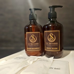 Dos botellas de champú y gel de baño Le Srivaree con dispensadores, sobre una superficie cubierta con un paño blanco y una bolsa de papel con la etiqueta 'SHOWER BAG'.