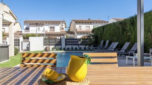 Zona junto a la piscina con una mesa de madera, sillas de descanso y vista despejada a un edificio residencial al fondo.