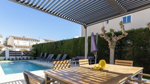 Zona junto a la piscina con mesa y sillas de madera, un área de estar cubierta y vista a un edificio residencial al fondo.