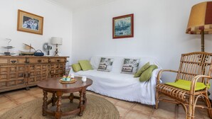 A cozy living room with a wooden cabinet, a round wooden table, a white sofa with green cushions, and a wicker chair.