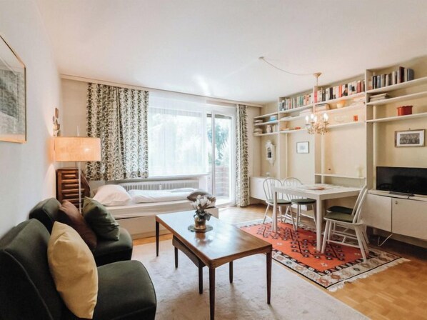 A cozy living room with a sofa, a coffee table, a dining area with chairs, a bookshelf, and a television.