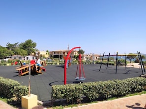 Ein Spielplatz im Freien mit Schaukeln und einer Wippe, umgeben von ordentlich geschnittenen Hecken und wolkenlosem Himmel.
