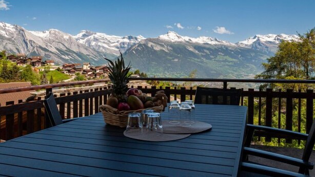 A table with a fruit basket, glasses, and a placemat, overlooking a mountain landscape with houses.