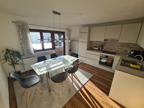 Une cuisine moderne avec des meubles blancs, un four encastré et un évier. On y trouve un coin repas avec une table et des chaises, ainsi qu’une plante en pot.